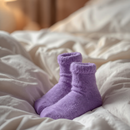 NINHO — Meias de dormir lavanda sobre cama com lençóis creme